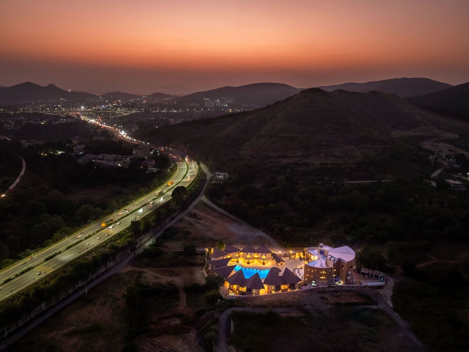 An aerial view of The Qualia Resort Club & Brewery, Udaipur at sunset, with mountains in the background and a road leading to the resort.