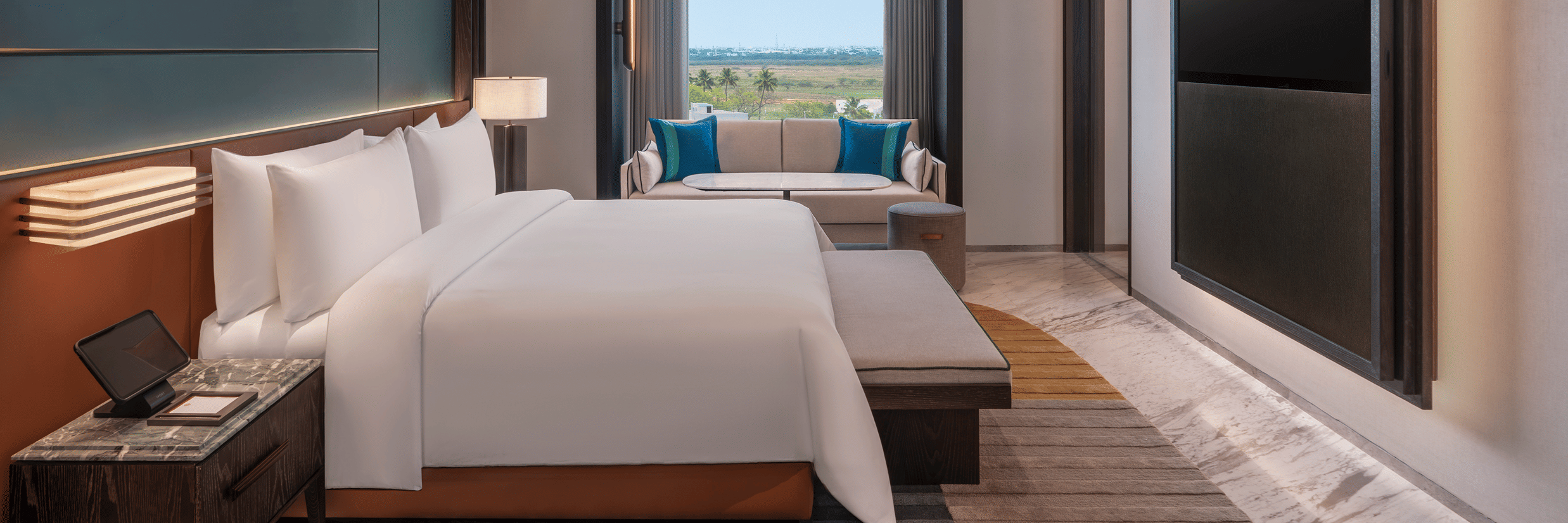 View of the Premium Suite bedroom at The Residency Signature Madurai with a king bed, wall-mounted TV, large window, and warm ambient lighting.