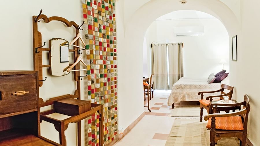 Hotel suite with a dressing area featuring a mirror, coat hooks, and a colorful hanging screen, leading to a bedroom with a bed and chairs through an arched opening at Tijara Fort-Palace, Alwar.