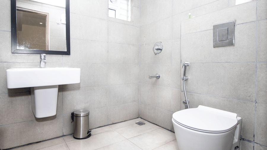 A bathroom at Sanctum Suites, Richmond Road, Bangalore features a pedestal sink, a metal bin, a toilet, a mirror, and tiled walls with chrome fixtures.