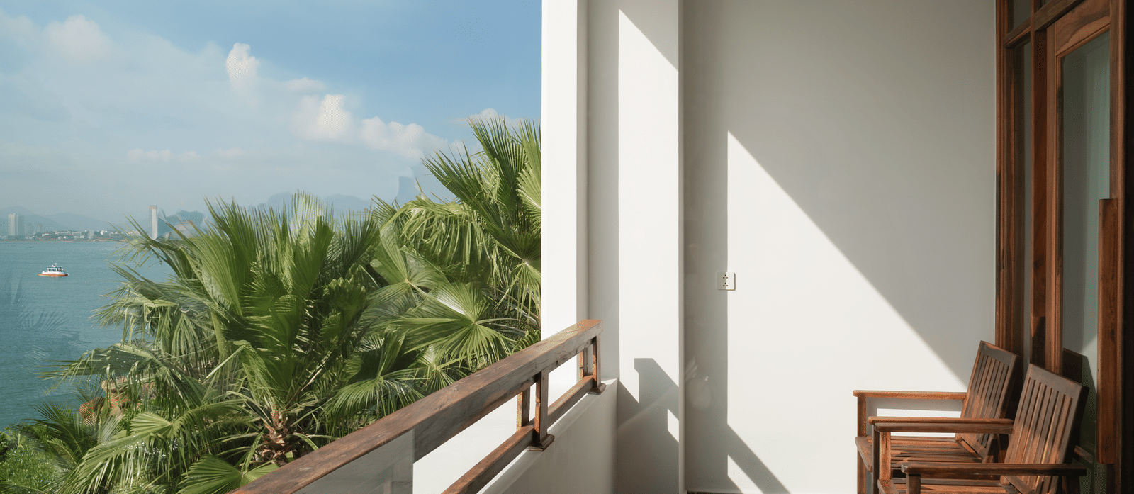 Private balcony at Alibu Resort Nha Trang, Vietnam, featuring wooden chair, tiled floor and glass balustrade overlooking palm trees and landscaped tropical gardens.