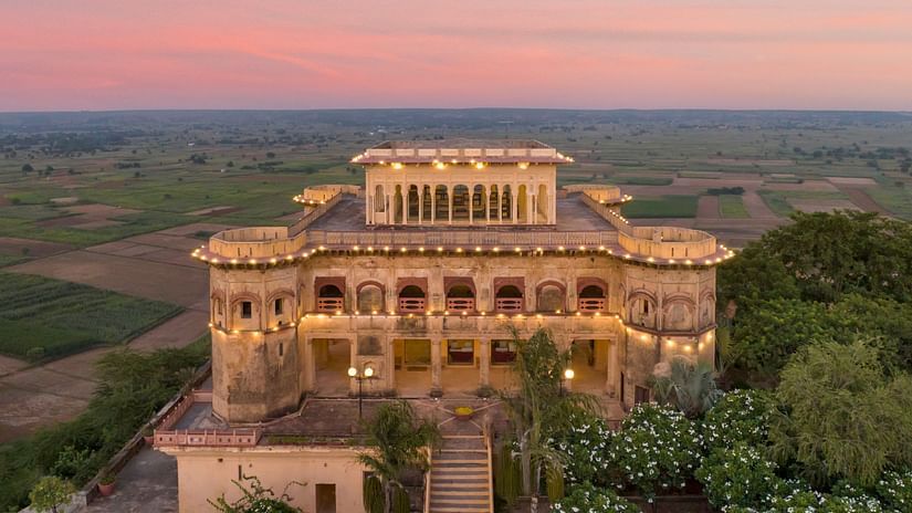 Facade image of Tijara Fort-Palace - 19th Century, Alwar surrounded by lush greenery with orange hued sky in the background.