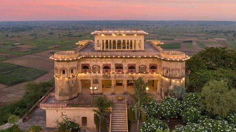 Facade image of Tijara Fort-Palace - 19th Century, Alwar surrounded by lush greenery with orange hued sky in the background.