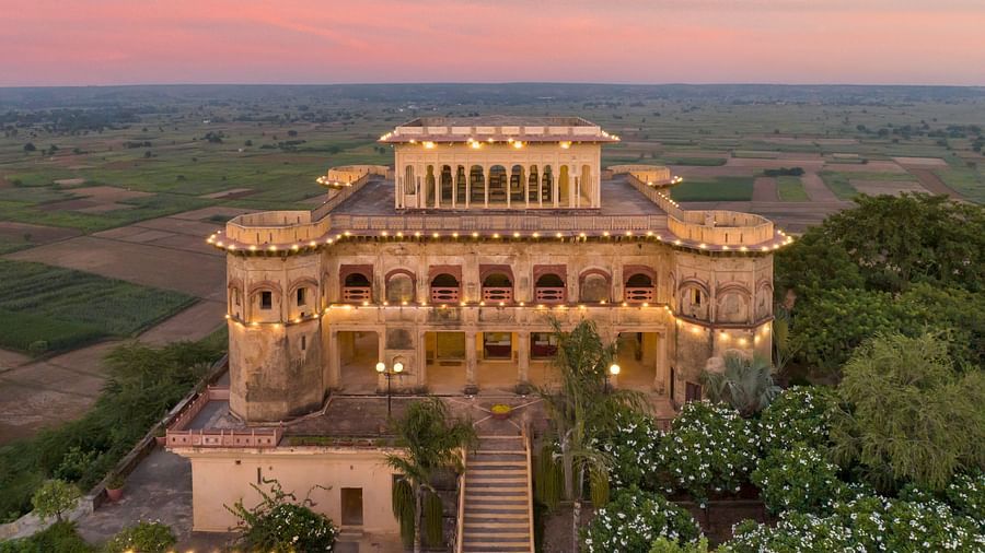 Facade image of Tijara Fort-Palace - 19th Century, Alwar surrounded by lush greenery with orange hued sky in the background.