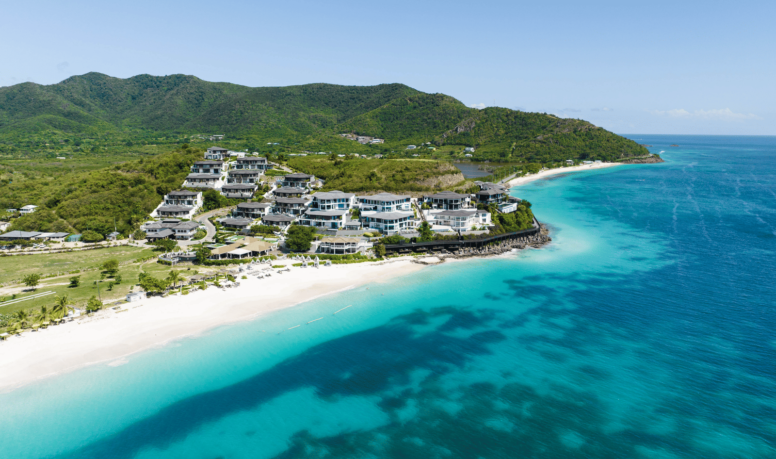 An aerial view of Tamarind Hills Resort and Villas - Antigua, nestled between lush green hills, sands and the sea.