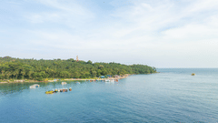 A tranquil Andaman coastline with calm blue waters, anchored boats, dense tropical greenery, and a distant lighthouse beneath a wide, cloud-streaked sky