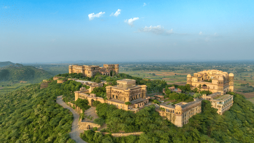 An aerial view of Tijara Fort-Palace atop a lush green hill, showcasing the fort's architecture and the surrounding landscape under a clear blue sky - Tijara Fort-Palace - 19th Century, Alwar.