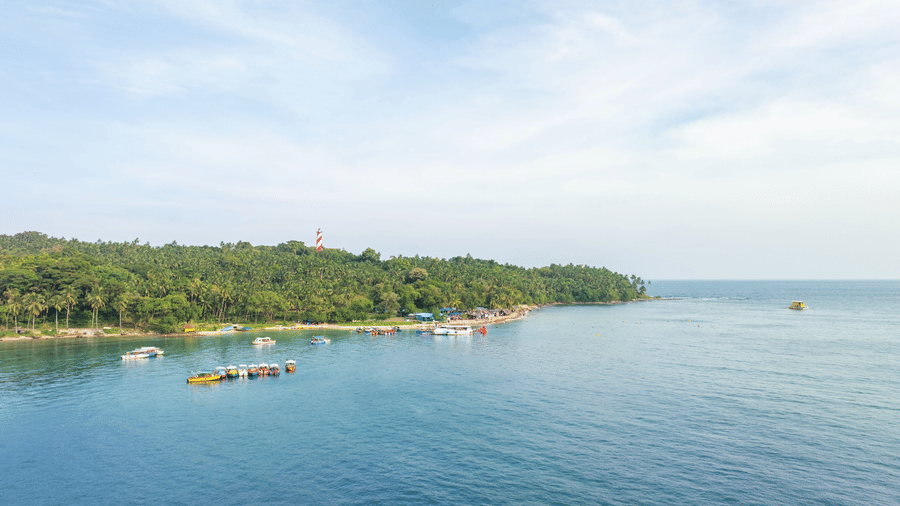 A tranquil Andaman coastline with calm blue waters, anchored boats, dense tropical greenery, and a distant lighthouse beneath a wide, cloud-streaked sky
