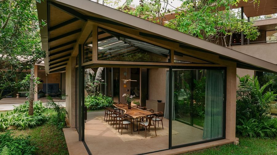 A well ventilated sun room with tall glass windows at Villa Moira by V Escapes, revealing a dining area surrounded by green lawns and vibrant plants.