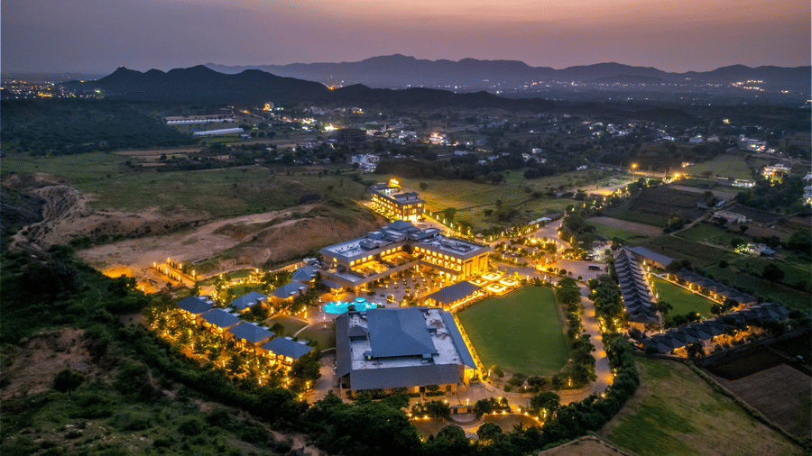 A birds eye view of the warmly lit Pushkara Resort & Spa, Ajmer, featuring accommodation blocks, a lawn, a pool, and sand dunes.