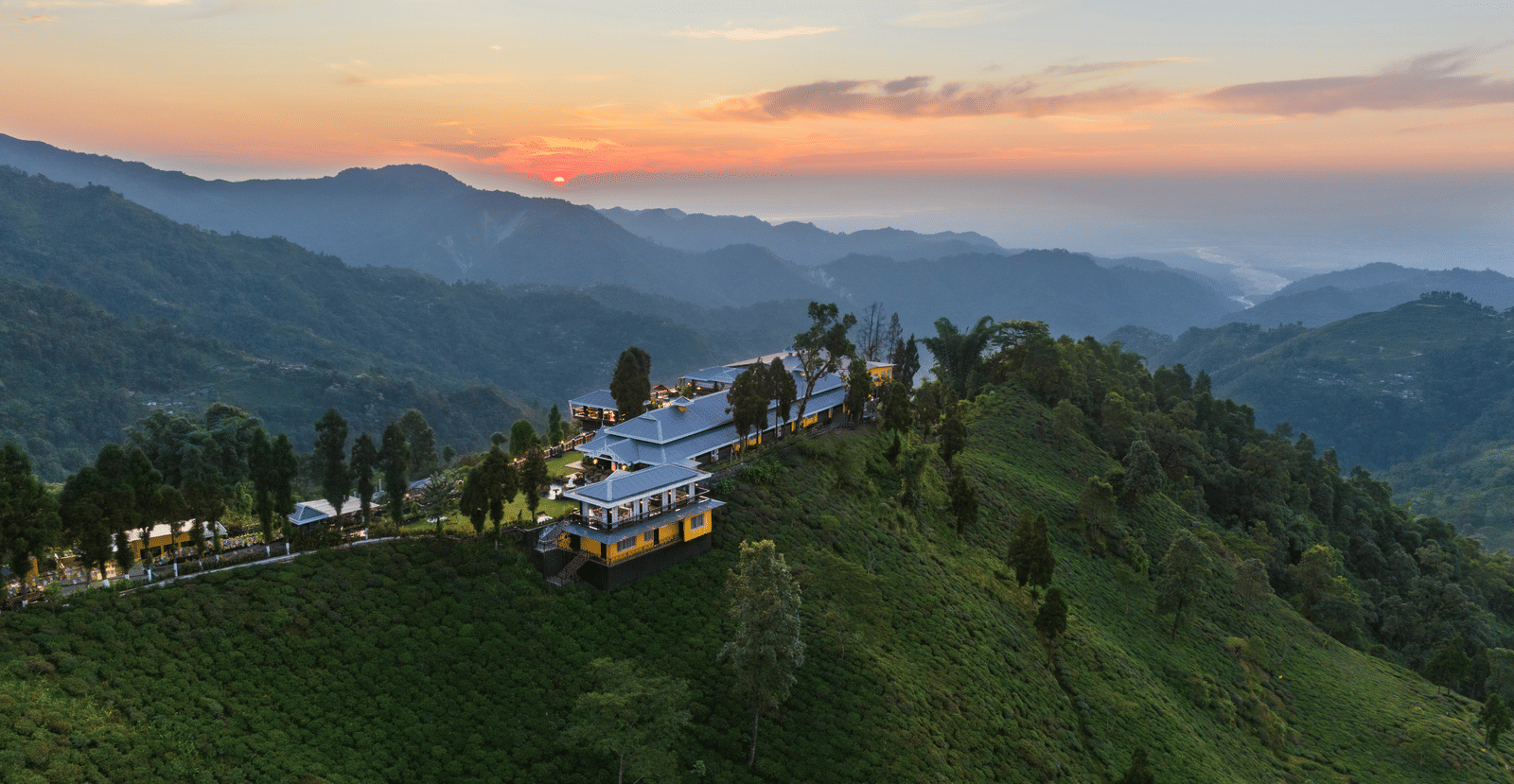 Aerial view of MAYFAIR Manor Jungpana featuring the resort building perched on a sloping green hill with distant mountains in the background and a setting sun. 