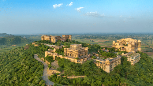 An aerial view of Tijara Fort-Palace atop a lush green hill, showcasing the fort's architecture and the surrounding landscape under a clear blue sky