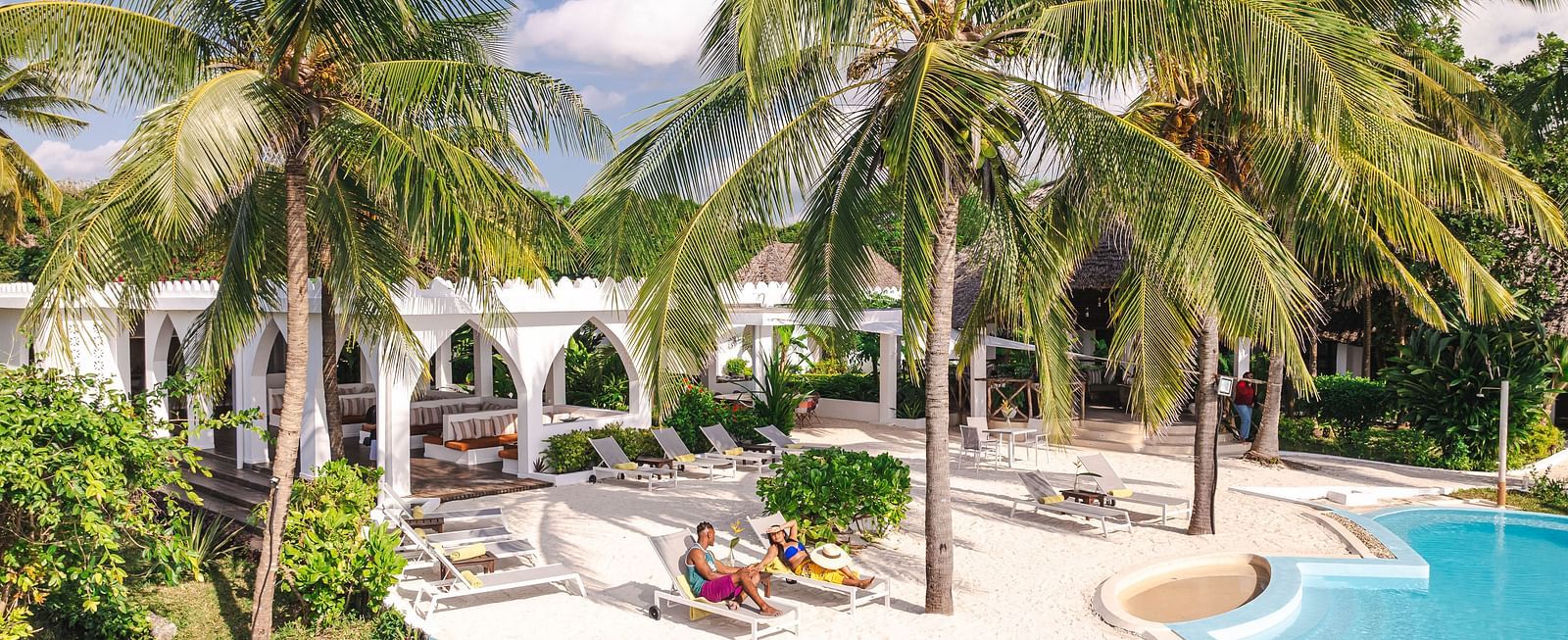 View of a swimming pool and a man and a woman with palm trees and buildings in the background - Shooting Star Boutique Hotel.