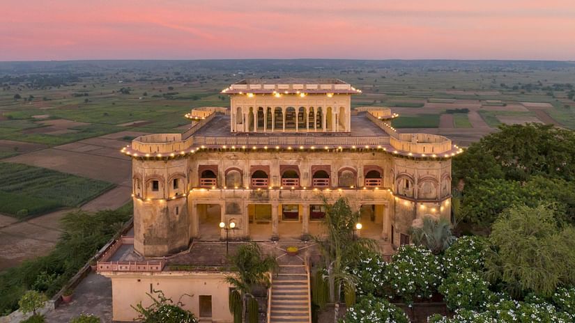 Facade image of Tijara Fort-Palace - 19th Century, Alwar surrounded by lush greenery with orange hued sky in the background.