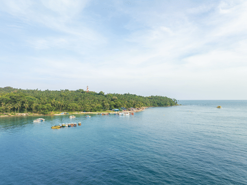 A tranquil Andaman coastline with calm blue waters, anchored boats, dense tropical greenery, and a distant lighthouse beneath a wide, cloud-streaked sky