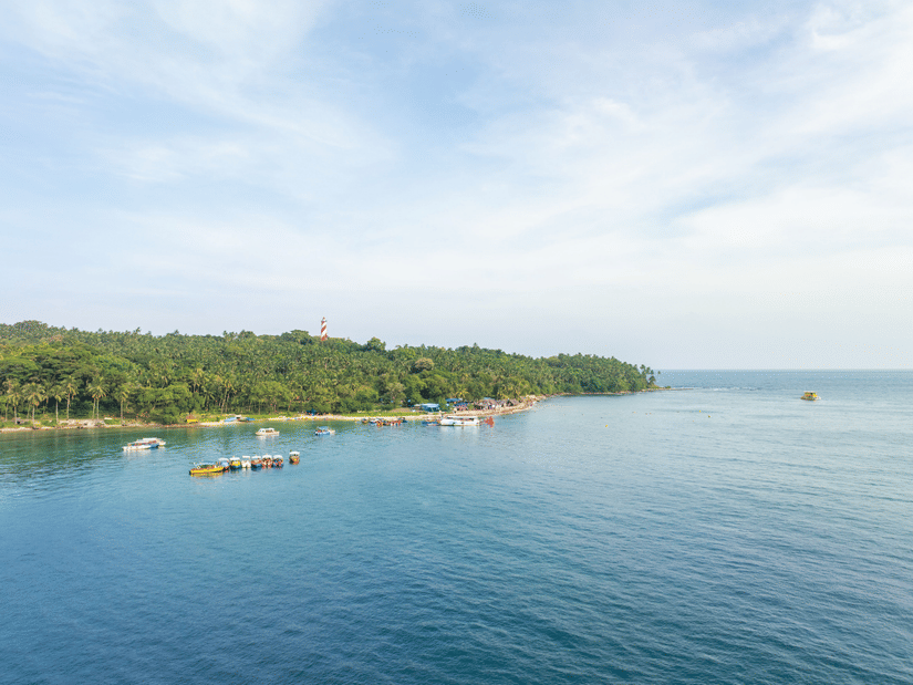 A tranquil Andaman coastline with calm blue waters, anchored boats, dense tropical greenery, and a distant lighthouse beneath a wide, cloud-streaked sky
