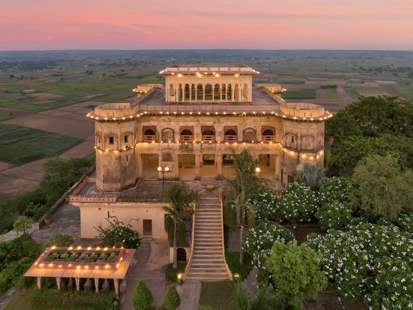 Facade image of Tijara Fort-Palace - 19th Century, Alwar surrounded by lush greenery with orange hued sky in the background. 