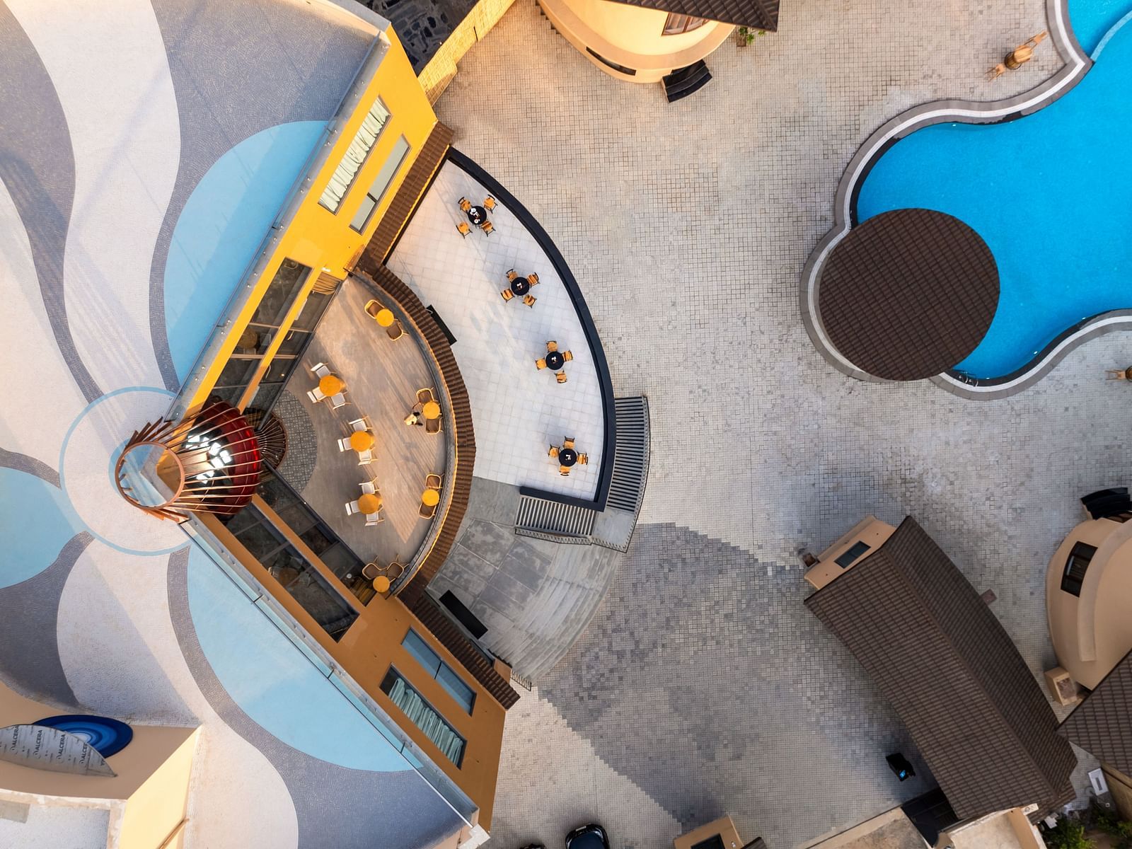 An aerial view of The Qualia Resort Club & Brewery, Udaipur capturing a uniquely designed pool area with geometric shapes and surrounding landscaping.
