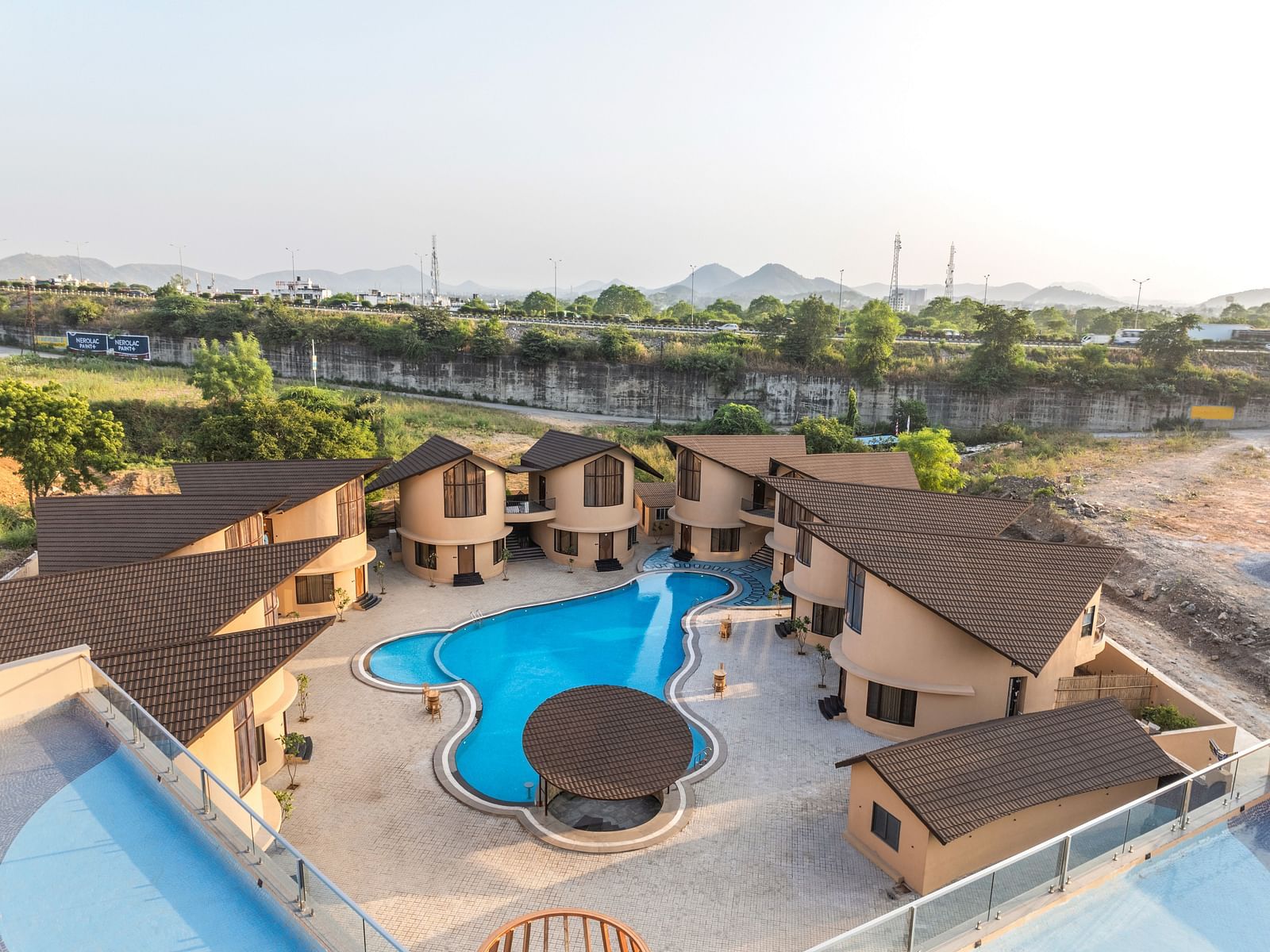 An overview of The Qualia Resort Club & Brewery, Udaipur with a central pool, surrounded by guest houses and natural landscape.