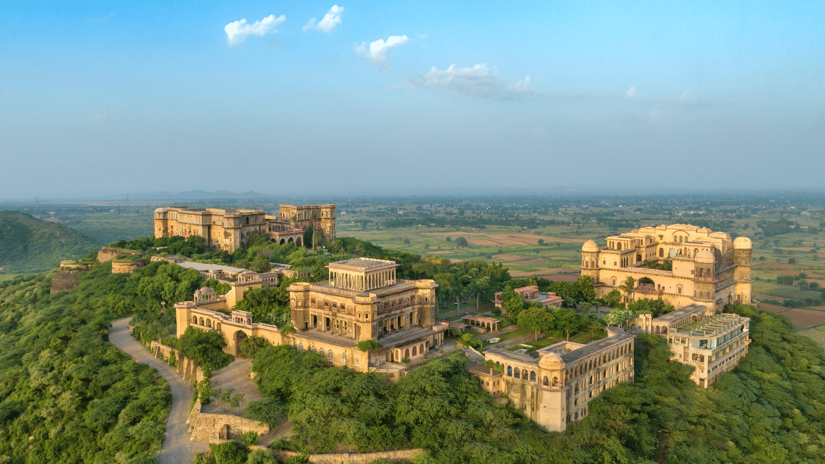 An aerial view of Tijara Fort-Palace atop a lush green hill, showcasing the fort's architecture and the surrounding landscape under a clear blue sky - Tijara Fort-Palace - 19th Century, Alwar.
