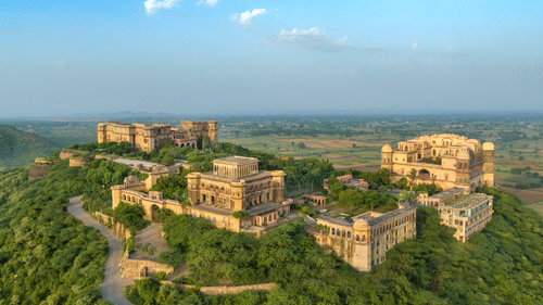 An aerial view of Tijara Fort-Palace atop a lush green hill, showcasing the fort's architecture and the surrounding landscape under a clear blue sky