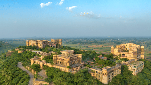 An aerial view of Tijara Fort-Palace atop a lush green hill, showcasing the fort's architecture and the surrounding landscape under a clear blue sky