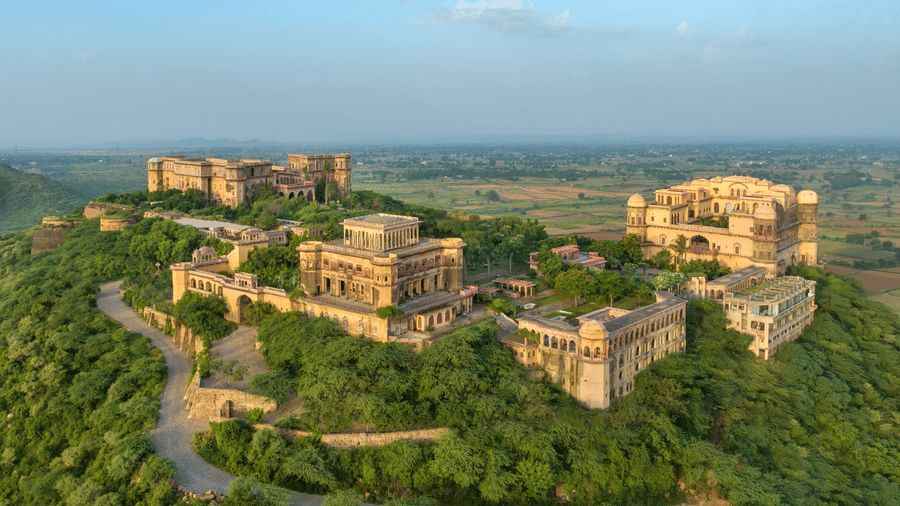 An aerial view of Tijara Fort-Palace atop a lush green hill, showcasing the fort's architecture and the surrounding landscape under a clear blue sky