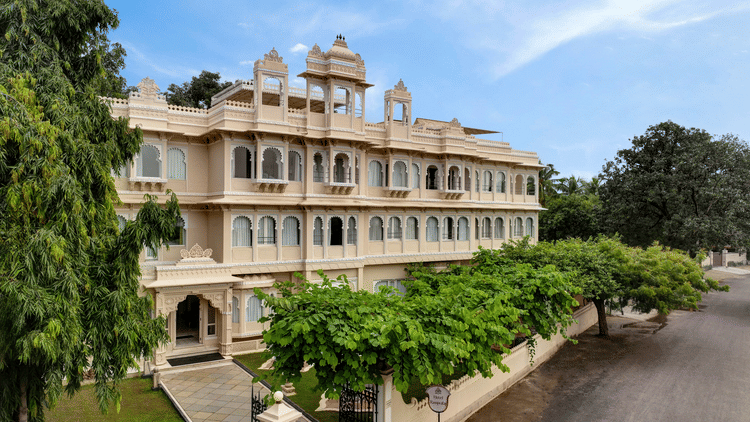 Front exterior of Ram Pratap Palace in Udaipur, India, with a large, traditional hotel building, green trees, and manicured landscaping under a blue sky.