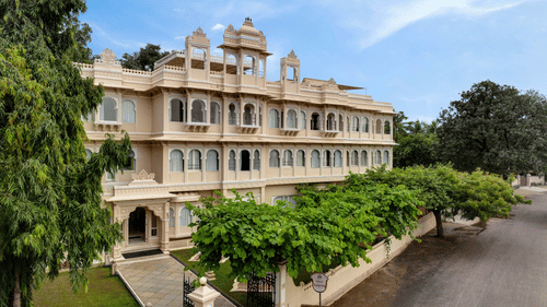 Front exterior of Ram Pratap Palace in Udaipur, India, with a large, traditional hotel building, green trees, and manicured landscaping under a blue sky.