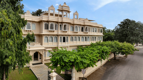 Front exterior of Ram Pratap Palace in Udaipur, India, with a large, traditional hotel building, green trees, and manicured landscaping under a blue sky.