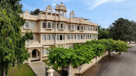 Front exterior of Ram Pratap Palace in Udaipur, India, with a large, traditional hotel building, green trees, and manicured landscaping under a blue sky.