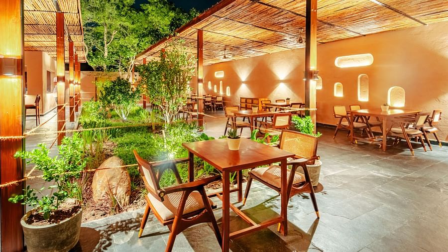 An image showing wooden chairs and tables arranged for a restaurant in an Eco-friendly theme, around green bushes, filled with bright yellow lights for the ambience.
