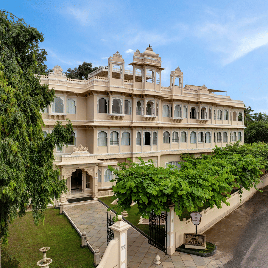 Front exterior of Ram Pratap Palace in Udaipur, India, with a large, traditional hotel building, green trees, and manicured landscaping under a blue sky.