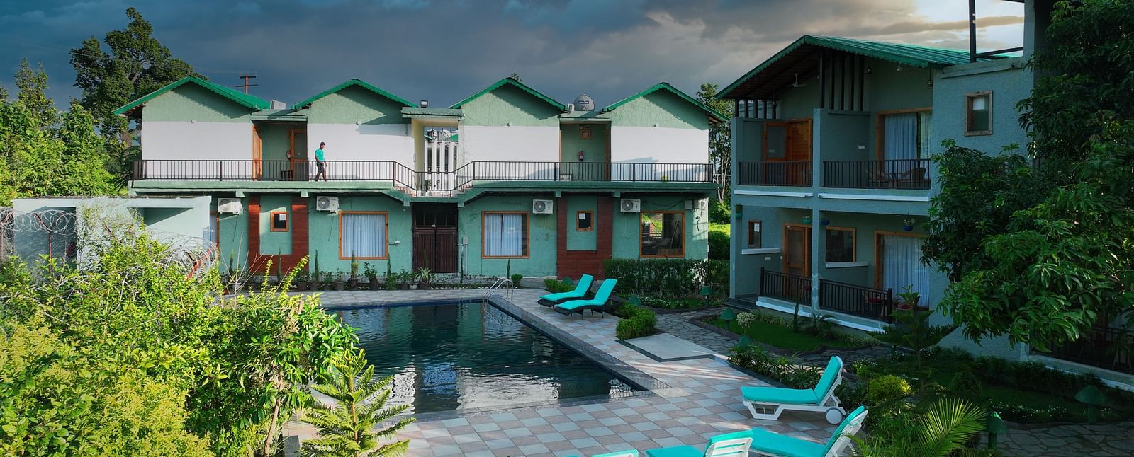 A resort complex with multiple buildings facing a swimming pool, surrounded by lush greenery under a dramatic sky.
