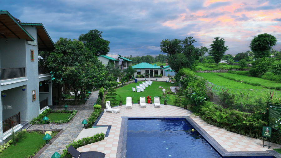 A top angle shot of the swimming pool sharing the grounds with an expansive green lawn under a picturesque sky at Corbett Nirvana Resort.