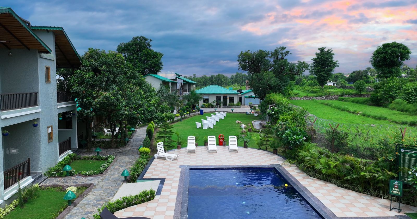 A top angle shot of the swimming pool sharing the grounds with an expansive green lawn under a picturesque sky at Corbett Nirvana Resort.