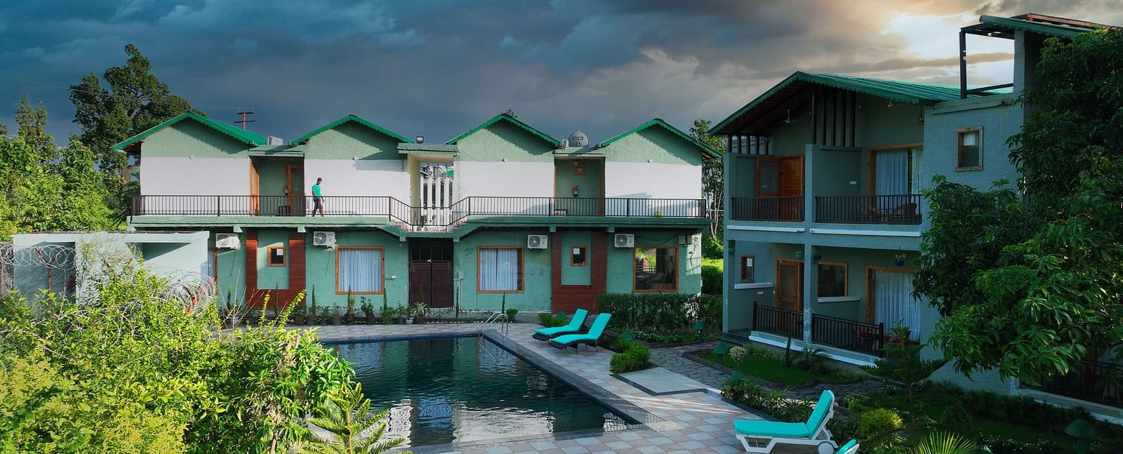A resort complex with multiple buildings facing a swimming pool, surrounded by lush greenery under a dramatic sky.