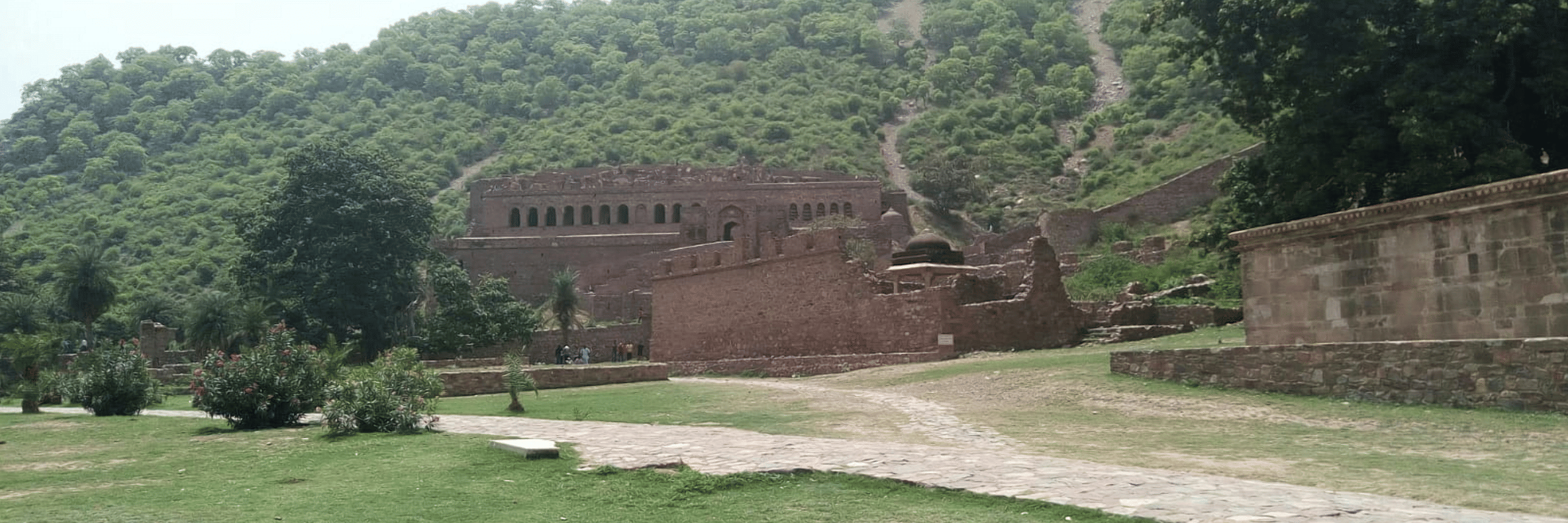 A view from far out of Bhangarh Fort, Alwar, with a pathway leading to the fort, a large lawn area, trees next to the fort and a mountain in the background.