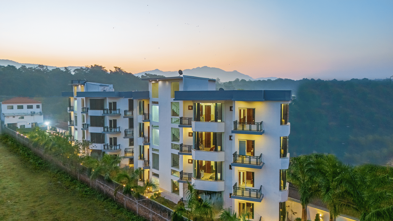 An aerial dusk view of a multi-storey white hotel building nestled against a lush green hillside - Nature Trails Rishikesh