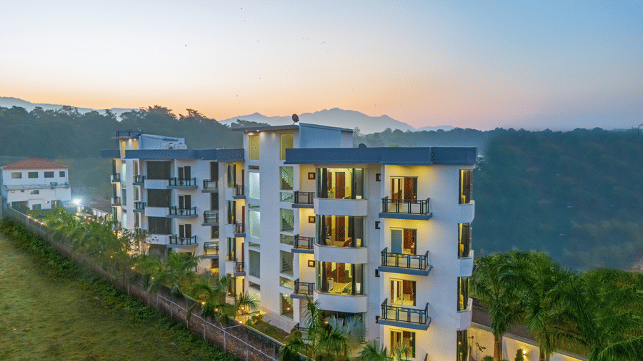 An aerial dusk view of a multi-storey white hotel building nestled against a lush green hillside - Nature Trails Rishikesh