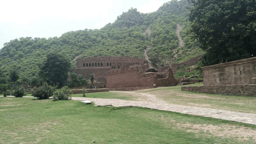 A view from far out of Bhangarh Fort, Alwar, with a pathway leading to the fort, a large lawn area, trees next to the fort and a mountain in the background.
