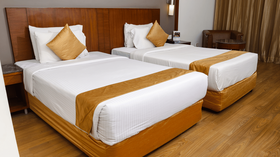 Twin bed room at Featherlite Evoma Hotel with beds dressed in white and gold linen, wooden headboards and long curtains by the window.
