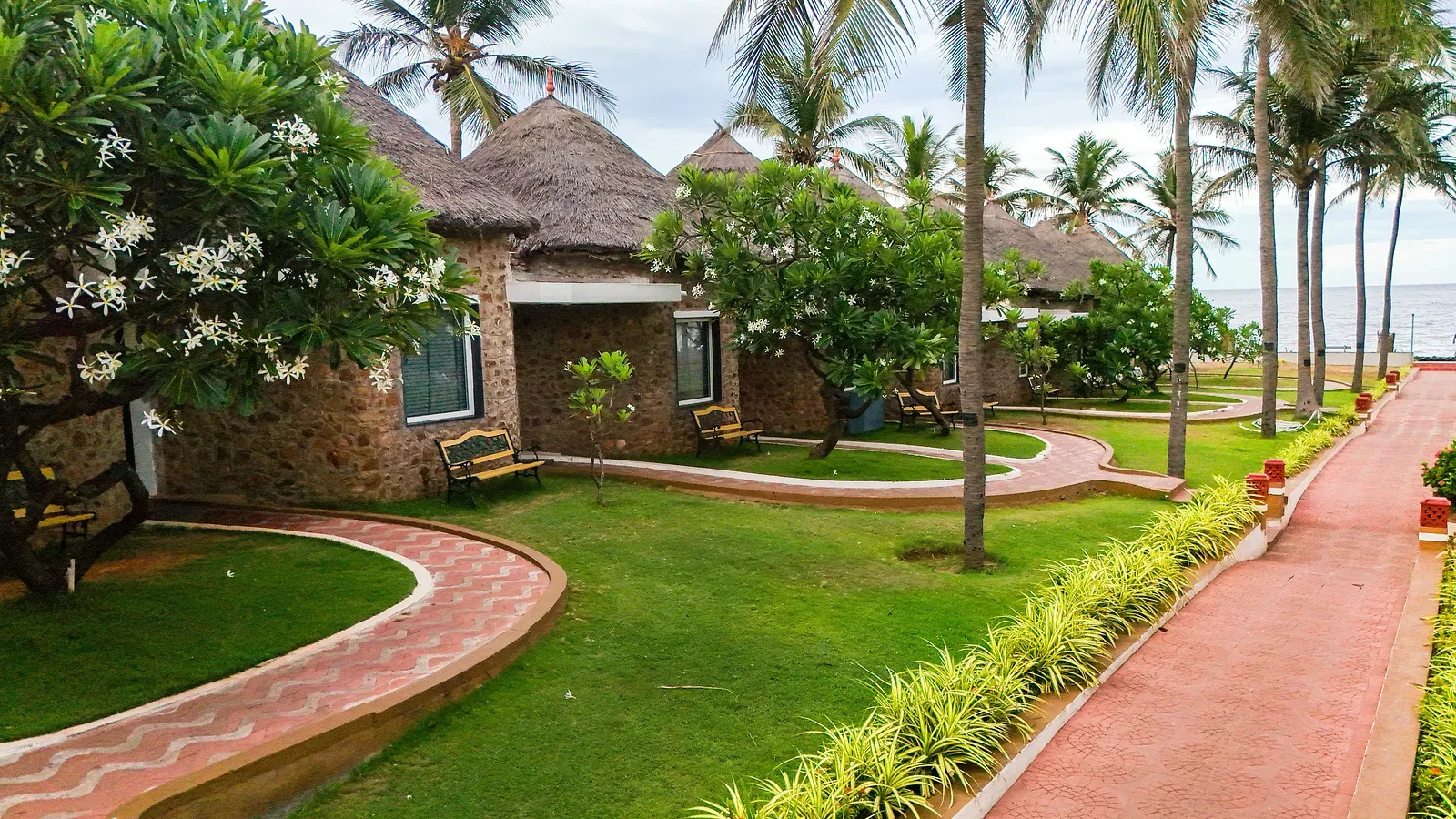 Villas near the beach with straw roofs and lawns in front, surrounded by coconut trees and tiled pathways leading from each villa