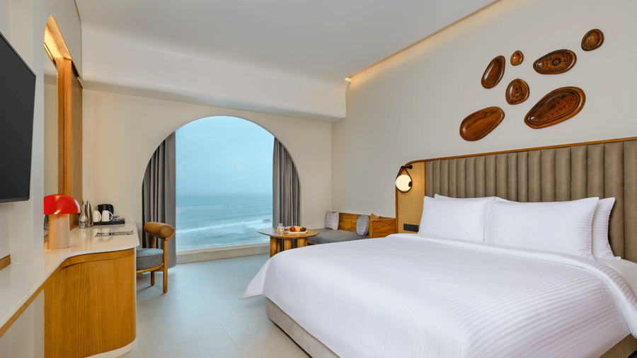 The Sea View Room (Front View) at Swosti Premium Beach Resorts, Puri, featuring a double bed, a television mounted on the wall, modern wooden decor, and large windows displaying a  sea view. 