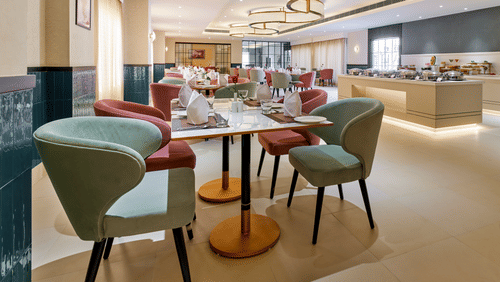 An image of a dining area featuring tables surrounded by chairs, with a buffet table elegantly set in the background at Tulip Inn, Bhopal.