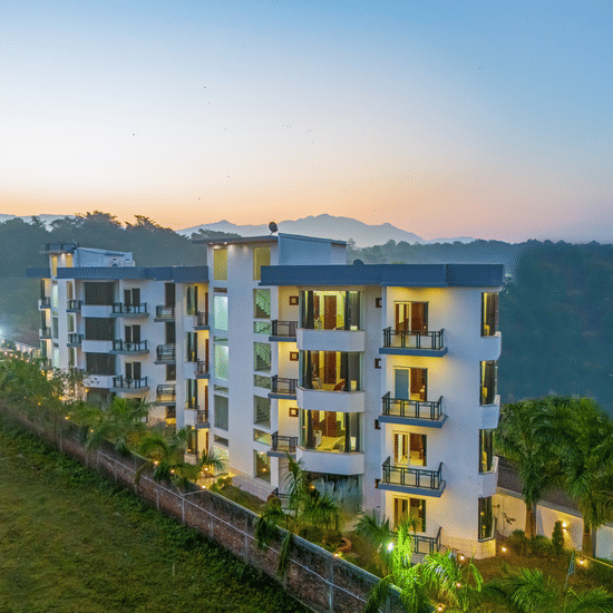 An aerial dusk view of a multi-storey white hotel building nestled against a lush green hillside - Nature Trails Rishikesh