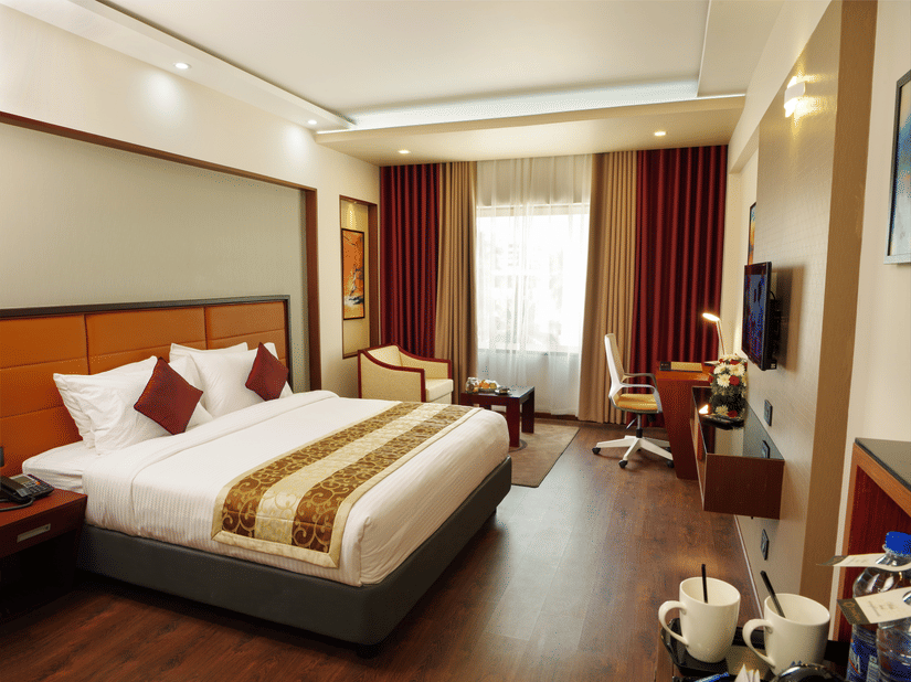 The Standard Room at Dimora, Thiruvananthapuram with a king size bed, a work desk, a flat screen television, a glass window with curtains, and wooden flooring.