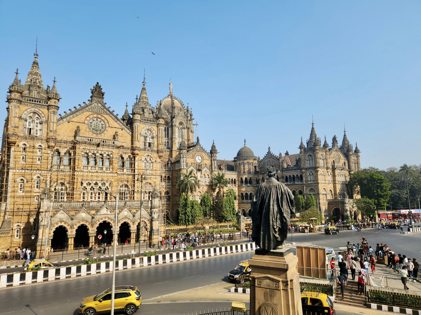 Chhatrapati Shivaji Maharaj Terminus in Mumbai with its historic Victorian Gothic architecture, a statue in the foreground, and a busy road with vehicles and pedestrians under a clear sky.