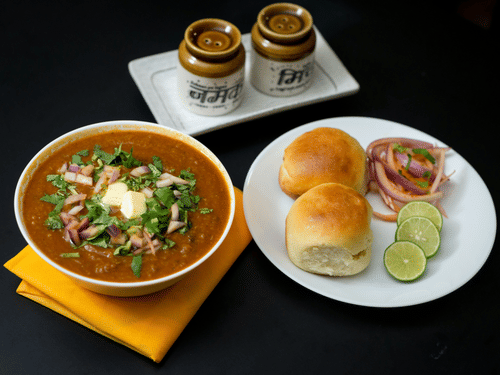 A plate filled with pav (bread), lime slices, and pickled onions, next to a big bowl with bhaji and 2 pickle jars nearby. 