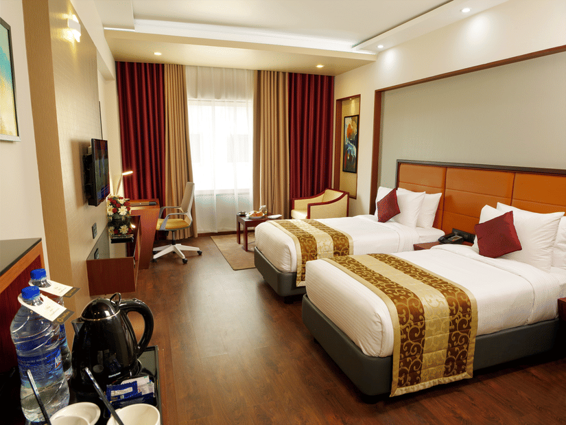 The Standard Room with twin beds at Dimora, Thiruvananthapuram, with twin beds, a flat screen television, glass window with curtains, and wodden flooring.